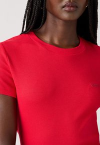 T-shirt rouge à rayures avec des manches courtes et un col rond. Présente une petite étiquette logo "Levi's" sur le côté inférieur. Texture de tissu en coton lisse.