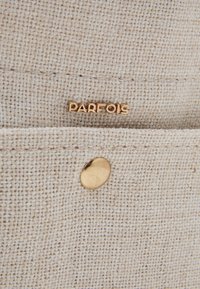 Beżowa tkanina o fakturowanej powierzchni, z złotym logo "PARFOIS" oraz okrągłym złotym zatrzaskiem.