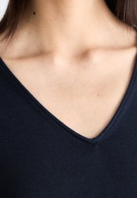 Młoda kobieta w granatowej bluzce z dekoltem w serek, odsłaniająca obojczyk i szyję, z gładką skórą i małą znamionem po lewej stronie.