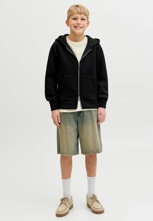 Smilende ung dreng iført en sort lynlås-hættetrøje, hvid skjorte, falmede denimbukser, hvide sokker og beige snøresko, stående på en ensfarvet baggrund.