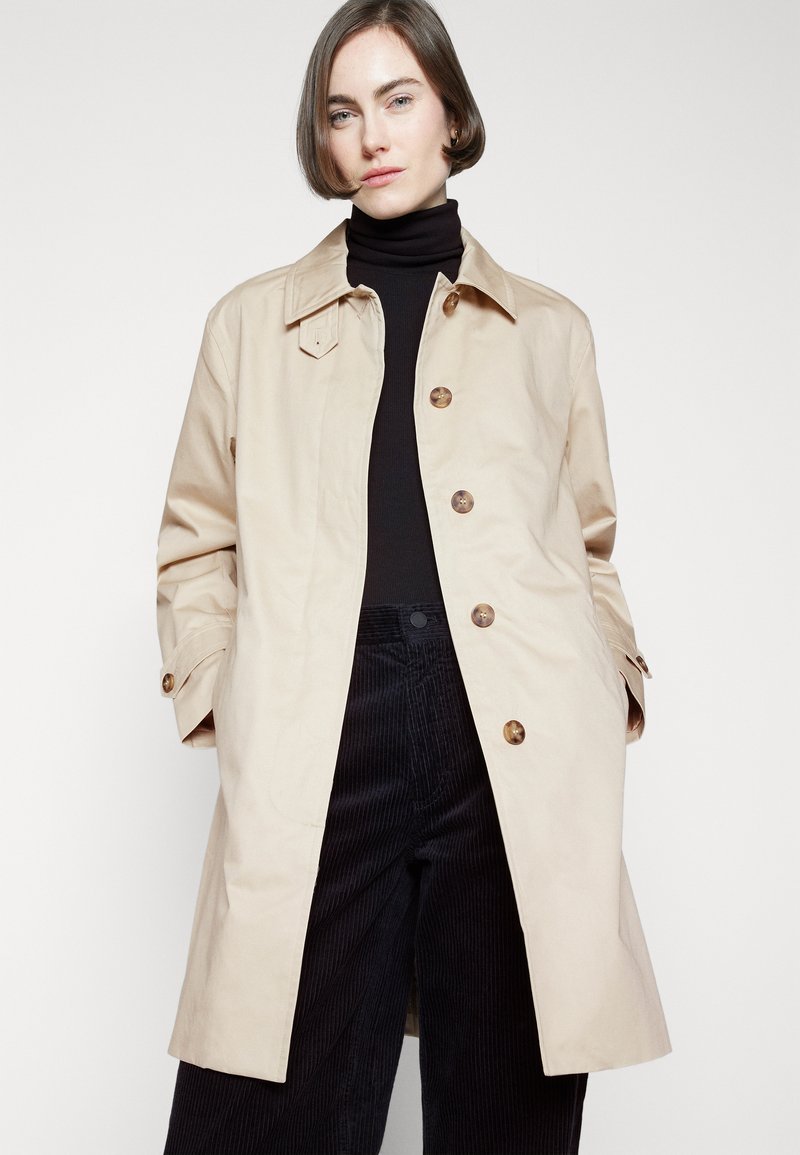 Abrigo beige claro con cuello, botones frontales y bolsillos laterales. Llevado sobre un suéter negro de cuello alto y pantalones de pana oscuros con rayas verticales.