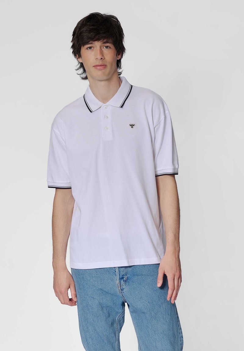 Ung mand iført en hvid polo-shirt med sort kant og et lille bi-logo, kombineret med lyseblå jeans, står foran en ensfarvet baggrund.