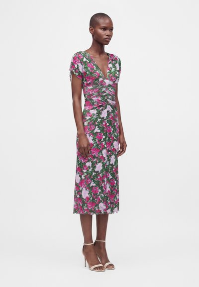 Femme portant une robe fleurie longueur genoux avec des motifs roses, violets et verts, devant froncé, manches courtes et sandales à talons hauts beiges.