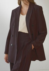 Blazer bordeaux croisé avec des boutons noirs, associé à un haut crème. Le tissu semble lisse avec des lignes structurées et une coupe ajustée.