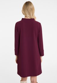 Woman with long blonde hair wears a long-sleeve, knee-length, plum-colored dress, standing facing away against a plain white background.