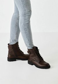 Stivaletti alla caviglia in finta pelle marrone con dettagli in lacci e cerniere, suole in gomma nera e finitura testurizzata, indossati con jeans azzurri chiari.