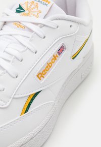 Närbild av vit Reebok Classic-sko med gula och gröna ränder, brittisk flagga-logotyp och "vegan"-etikett på tungan.