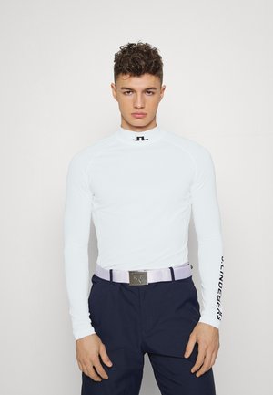 White long-sleeve athletic top with a high neck, black logo on front, and contrasting black text on the right sleeve. Paired with dark pants and a light belt.
