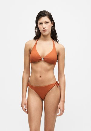Jeune femme aux cheveux mouillés portant un bikini texturé couleur rouille avec un détail anneau sur le haut et des liens latéraux sur le bas.