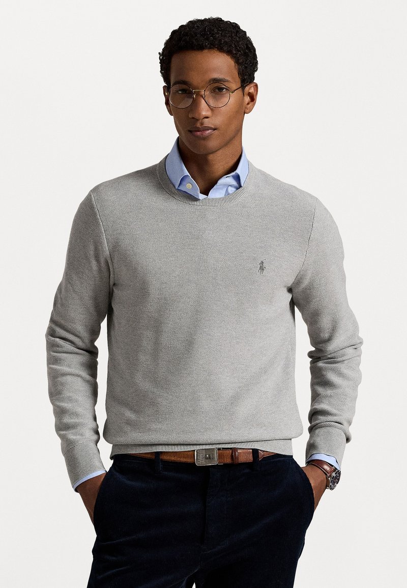 Lys grå strikket genser med rund hals, ribbestrikkede mansjetter og kant. Bæres over en lys blå skjorte med krage. Små logo detaljer til stede.