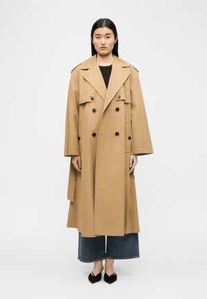 Egyenes fekete hajú nő bézs kétsoros gombú ballonkabátot, széles szárú, rövid farmernadrágot és fekete hegyes orrú cipőt visel, fehér háttér előtt áll.