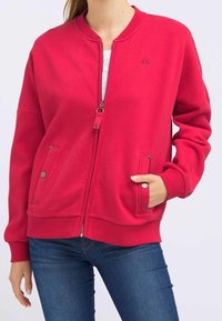 Veste zippée rouge avec col côtelé, deux poches avant à fermeture à pression, et coupe ajustée. Tissu en coton lisse avec un détail logo.