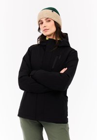 Vrouw in zwarte hoodie en groen-beige gebreide muts, staand met armen gekruist tegen een witte achtergrond.