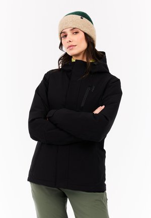 Vrouw in zwarte hoodie en groen-beige gebreide muts, staand met armen gekruist tegen een witte achtergrond.