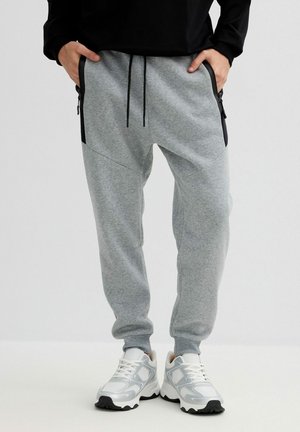 Persona con pantalones jogger gris claro con bolsillos con cremallera negra y zapatillas blancas, de pie con las manos en los bolsillos contra un fondo liso.