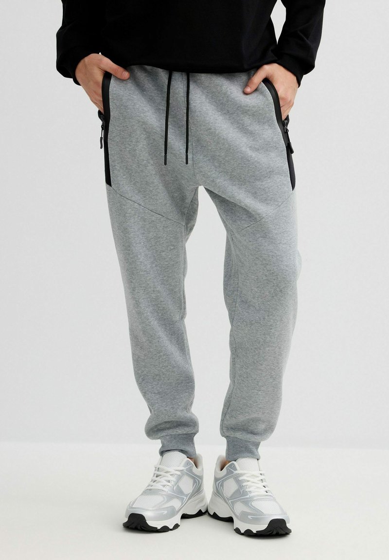 Persona con pantalones jogger gris claro con bolsillos con cremallera negra y zapatillas blancas, de pie con las manos en los bolsillos contra un fondo liso.