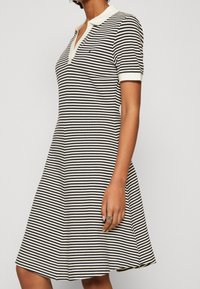 Pruhované polo šaty v černé a bílé, ribbed tkanina, krátké rukávy a výstřih do V s krémovým límcem a logem na levé straně hrudi.