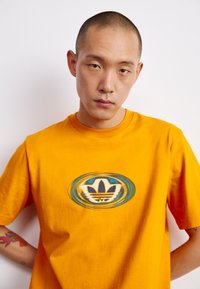 T-shirt di cotone arancione con un logo rotondo raffigurante un design a tre foglie in blu e bianco, maniche corte e vestibilità rilassata.