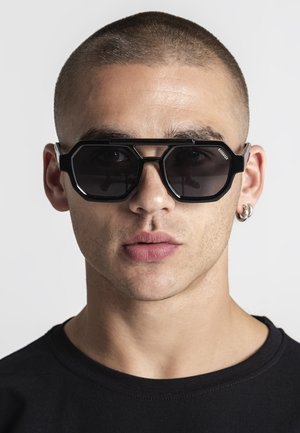 Mladý človek s krátkymi vlasmi, ktorý nosí veľké čierne geometrické slnečné okuliare a strieborné kruhové náušnice, oblečený v čiernej košeli, smerujúci vpred.