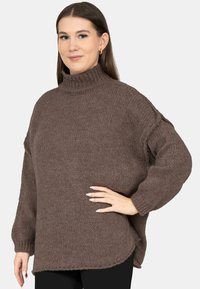 Vrouw met lang bruin haar, goudkleurige creolen en een losse, bruine gebreide coltrui, poserend met één hand op haar heup.