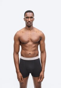 Svalnatý muž stojaci tvárou vpred, oblečený v čiernych boxerkách Calvin Klein, na jednoduchom bielom pozadí.