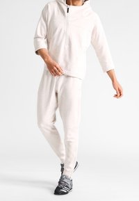 Sudadera ligera de color gris claro con capucha, cierre de cuarto de cremallera y mangas hasta tres cuartos, combinada con pantalones ajustados a juego y zapatillas deportivas negras.