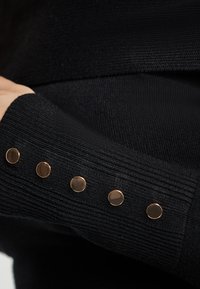 Pulover negru din tricot cu textura ribbed și accente metalice de culoare aurie pe manșetă, având cinci butoane decorative circulare.