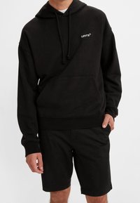 Haut à capuche Levi's noir avec cordons de serrage, poche kangourou et logo. Associé à un short noir, il présente une coupe décontractée et une texture douce.
