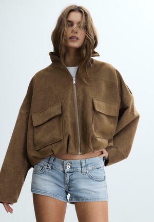 Ung kvinde iført en oversized brun ruskind jakke med lynlås og store lommer samt lyseblå denimshorts, stående foran en ensfarvet baggrund.