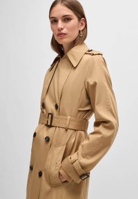 Beige Trenchcoat mit tailliertem Gürtel, großen Revers und Knopfdetails. Besitzt eine glatte Textur und lange Ärmel mit Schnallenakzenten.