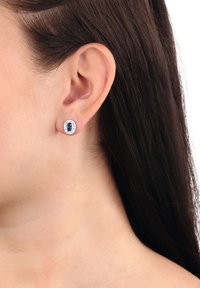 Kvinnas öra som bär en silverstud med en central mörkblå oval ädelsten omgiven av klara stenar.