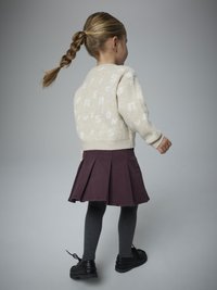 Kremowy sweter z białym napisem, w połączeniu z fioletową plisowaną spódnicą, szarymi prążkowanymi rajstopami i czarnymi butami. Widok od tyłu.