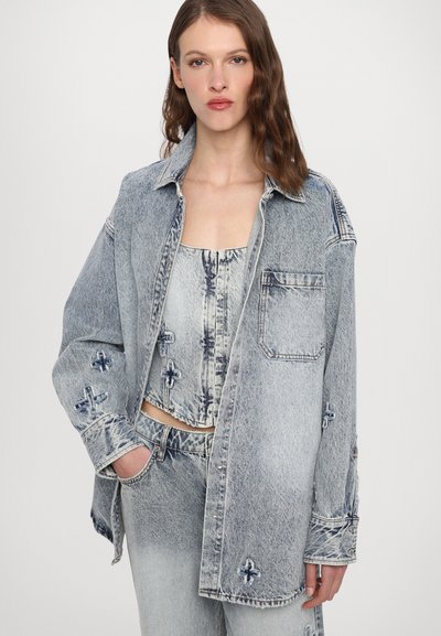 Šviesiai mėlynas džinsinis marškinis su laisvu kirpimu, matomomis siūlėmis ir aplikacijomis ant rankovių. Dėvimas ant atitinkamo trumpa viršaus.