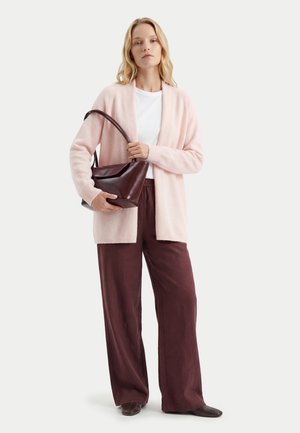 Frau trägt hellrosa Cardigan, weißes Top, weite braune Hose, hält dunkelbraune Lederhandtasche, eine Hand am Träger und die andere stützt die Basis.