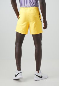 Gula sportshorts med elastisk midja, svarta sidokanter och ett lätt material, som är kombinerade med vita sneakers.