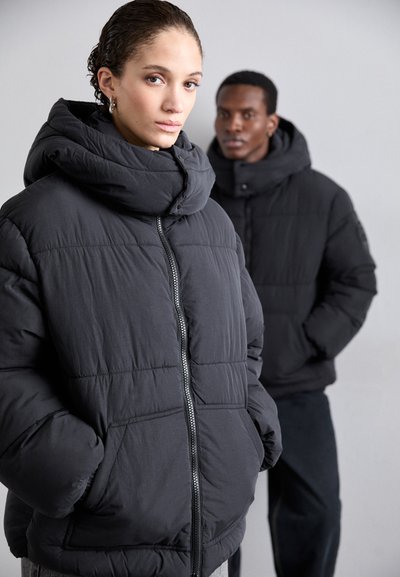 Black puffer jacket with a high collar, zipper closure, and quilted texture. Model features a relaxed fit and front patch pockets.