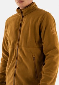 Chaqueta de forro polar marrón con cuello alto, cierre frontal con cremallera y bolsillo lateral con cremallera. Presenta un tejido texturizado y suave, y un detalle de logo en la manga.