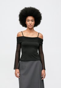 Femeie cu coafură afro, purtând un top negru cu umerii la vedere, cu mâneci lungi transparente și o fustă gri, stând în fața unui fundal simplu.