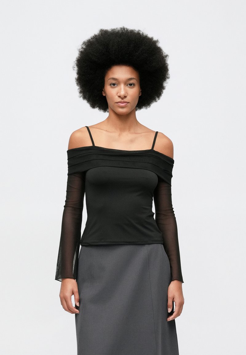 Femeie cu coafură afro, purtând un top negru cu umerii la vedere, cu mâneci lungi transparente și o fustă gri, stând în fața unui fundal simplu.