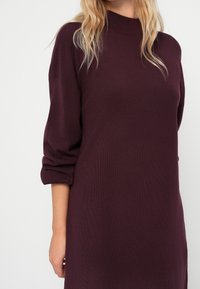 Robe en maille de couleur bordeaux foncé avec un col montant et des manches trois-quarts. Texture douce et coupe décontractée, avec des revers de manches retroussés.
