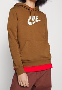 Brun huvtröja med en vit Nike-logotyp, snoddar vid huvan, en framficka, bärs över en röd tröja, ihop med bruna byxor.