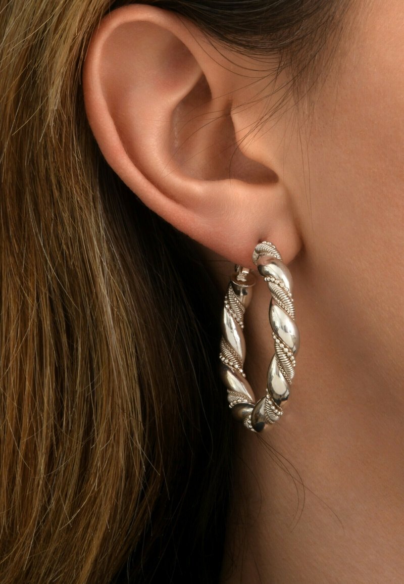 Les boucles d'oreilles en argent affichent un design torsadé avec des accents texturés et une finition polie. Portées à l'oreille, mettant en valeur les détails complexes.