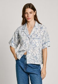 Kortermet skjorte i hvitt stoff med blå blomsterdesign, med en avslappet passform, krage og en enkelt brystlomme. Kombinert med blå jeans.