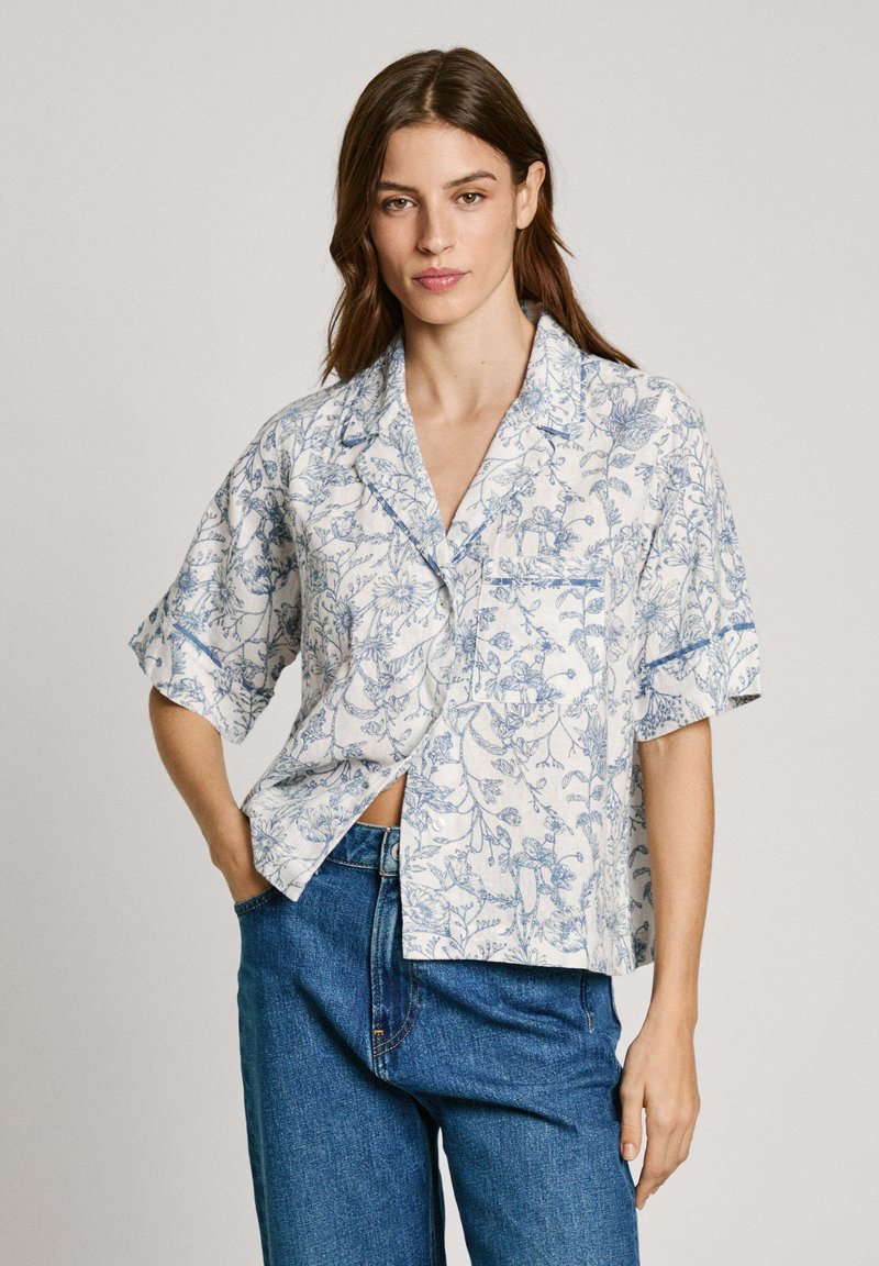 Kortermet skjorte i hvitt stoff med blå blomsterdesign, med en avslappet passform, krage og en enkelt brystlomme. Kombinert med blå jeans.