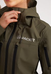 Giacca impermeabile verde oliva con colletto alto, cerniera frontale e dettagli neri. Presenta il logo del marchio "JACKIT" e due tasche con zip.