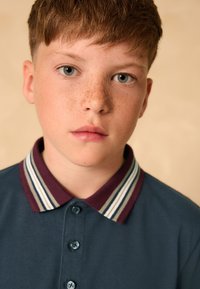 Marinblå pikétröja med randig vinröd och vit krage; har knäppning och mjuk textur; modellen har lätta fräknar och blå ögon.