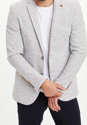 Homme portant un blazer gris clair texturé sur une chemise blanche avec un pantalon noir, les mains jointes devant, visible partiellement du cou aux cuisses.