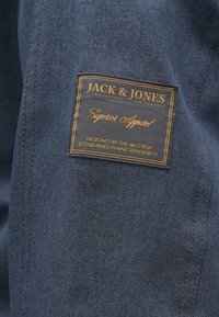 Giubbotto di jeans con un'etichetta in pelle marrone. L'etichetta presenta "Jack & Jones" in testo dorato e "Superior Apparel" in corsivo.