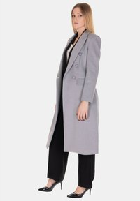 Cappotto doppiopetto grigio chiaro con vestibilità sagomata, dotato di rever a incavo e tasche laterali, abbinato a pantaloni neri a gamba larga e scarpe con punta affilata.