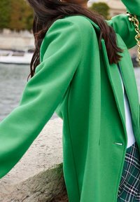 Zelený strukturovaný kabát s hladkým povrchem, dlouhými rukávy a jemným polstrováním na ramenou. Obsahuje štíhlou boční kapsu a kontrastní světlou vrstvu pod ním.